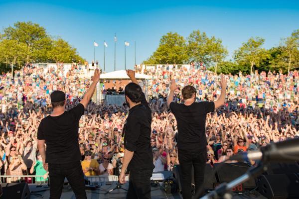 A band playing on stage - three men in black teeshirts - looking out into a large cloud on a warm, sunny day.