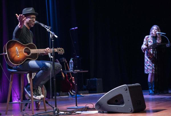 Keb’ Mo’ sitting on a stool, wearing blue jeans, a black shirt, and a black fedora hat. He has his guitar on his lap and is gesturing out to the audience. 