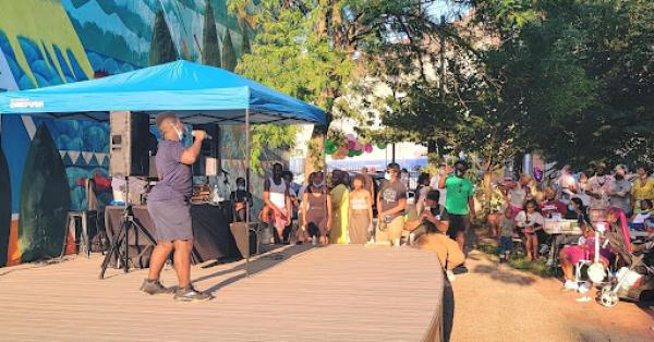 A Village Industries program in action. Someone is speaking passionately from the stage while a crowd of young people look on in brightly colored clothing - both standing and sitting. 