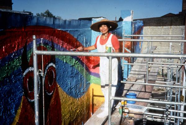 Iconic artist Lily Yeh painting wipe stripes of color on a mural. She is wearing white overalls and a sunhat. 