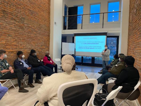 Community members gather at the Civic Power Studio. They are seated in a circle in folding chairs while watching a presentation.