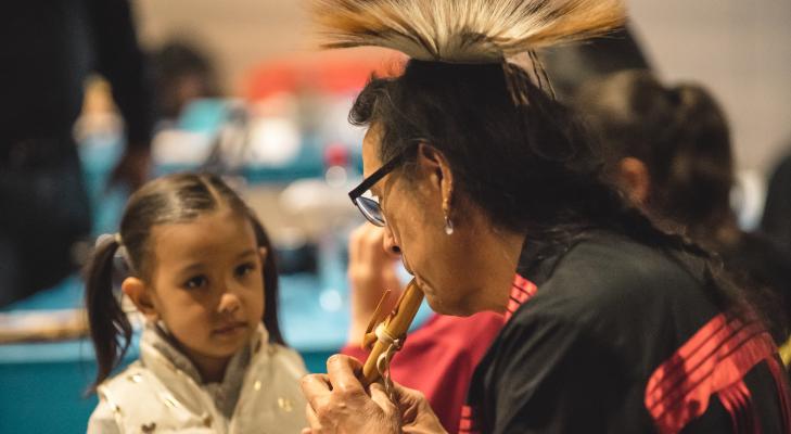 Indigenous artist, Tchin, plays a courting flute in front of a little girl with brown pigtails.