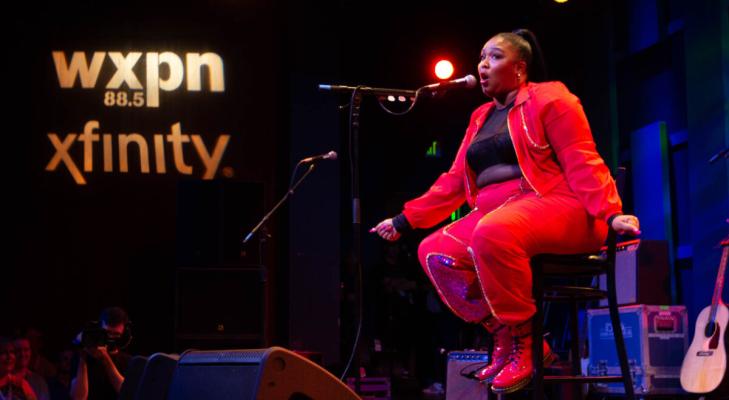 Lizzo sitting onstage at a WXPN show wearing a red track suit and her hair pulled back