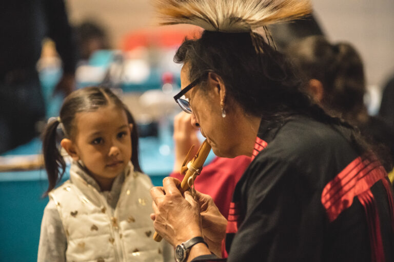 Indigenous artist, Tchin, plays a courting flute in front of a little girl with brown pigtails.