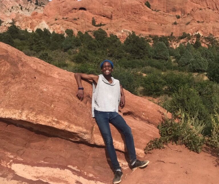 B.J. posing on a hill of orange rock. They are wearing a white top, jeans and a huge smile!