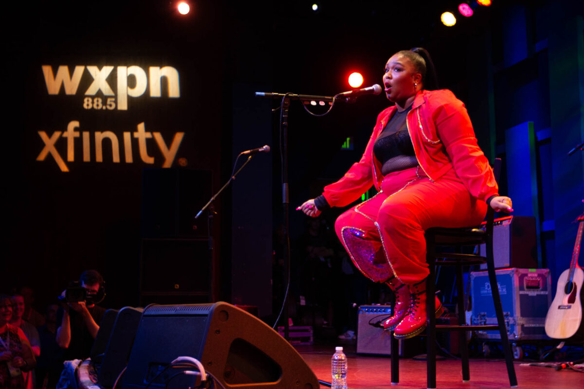 Lizzo sitting onstage at a WXPN show wearing a red track suit and her hair pulled back